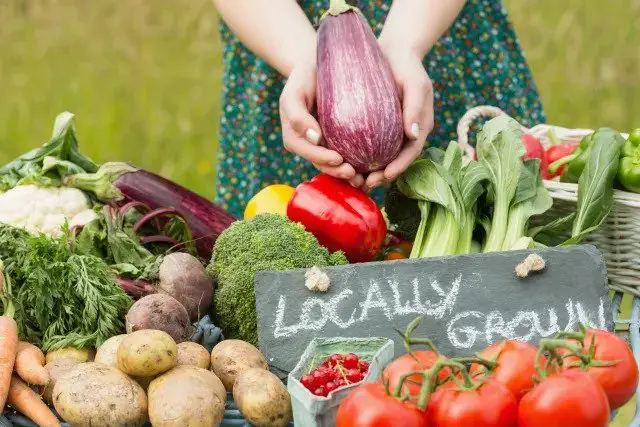 A woman is holding a basket of vegetables with a sign that says locally grown.