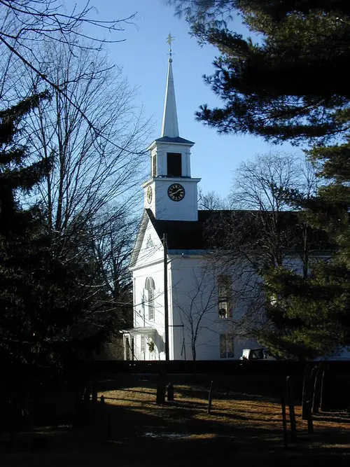 A white church with a steeple in the middle of a wooded area.